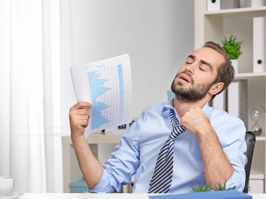 Gestresster Mann im Büro, der sich wegen Hitze Luft zufächelt – Symbolbild für schlechte Raumklimatisierung am Arbeitsplatz