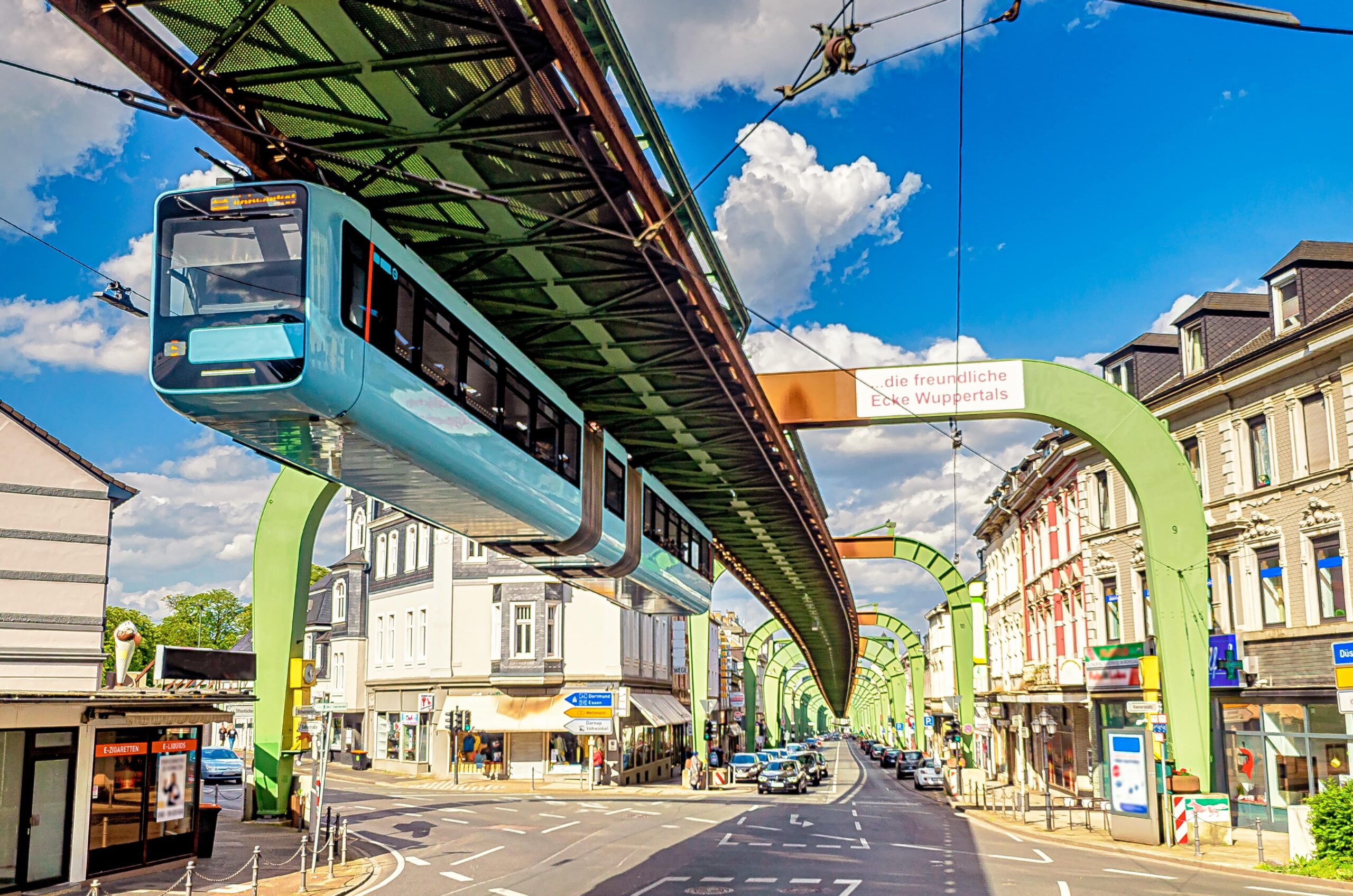 Schwebebahn in Wuppertal über der Stadtstraße, umgeben von bunten Gebäuden und blauem Himmel. Climate Energy.