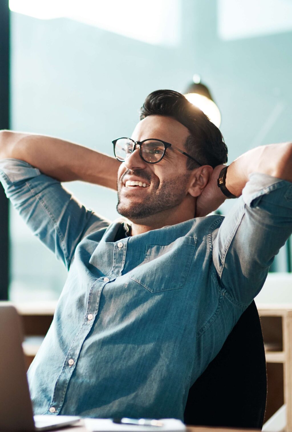Mann mit Brille entspannt am Schreibtisch, lächelnd, mit Laptop. Fokus auf positive Arbeitsatmosphäre. Climate Energy.