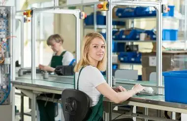 Eine junge Frau in Arbeitskleidung sitzt an einem Tisch in einer Produktionsumgebung. Climate Energy.