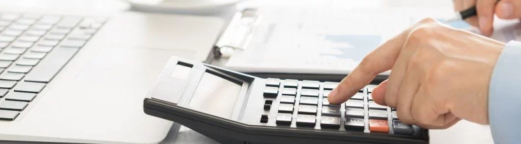 Hände tippen auf einem Taschenrechner neben einem Laptop und Dokumenten. Thema: Climate Energy.