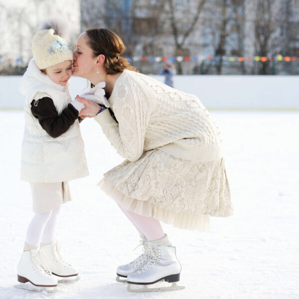 Eine Mutter küsst liebevoll ihre Tochter auf einer Eisbahn. Winterkleidung und Schlittschuhe betonen die Szene. Climate Energy.