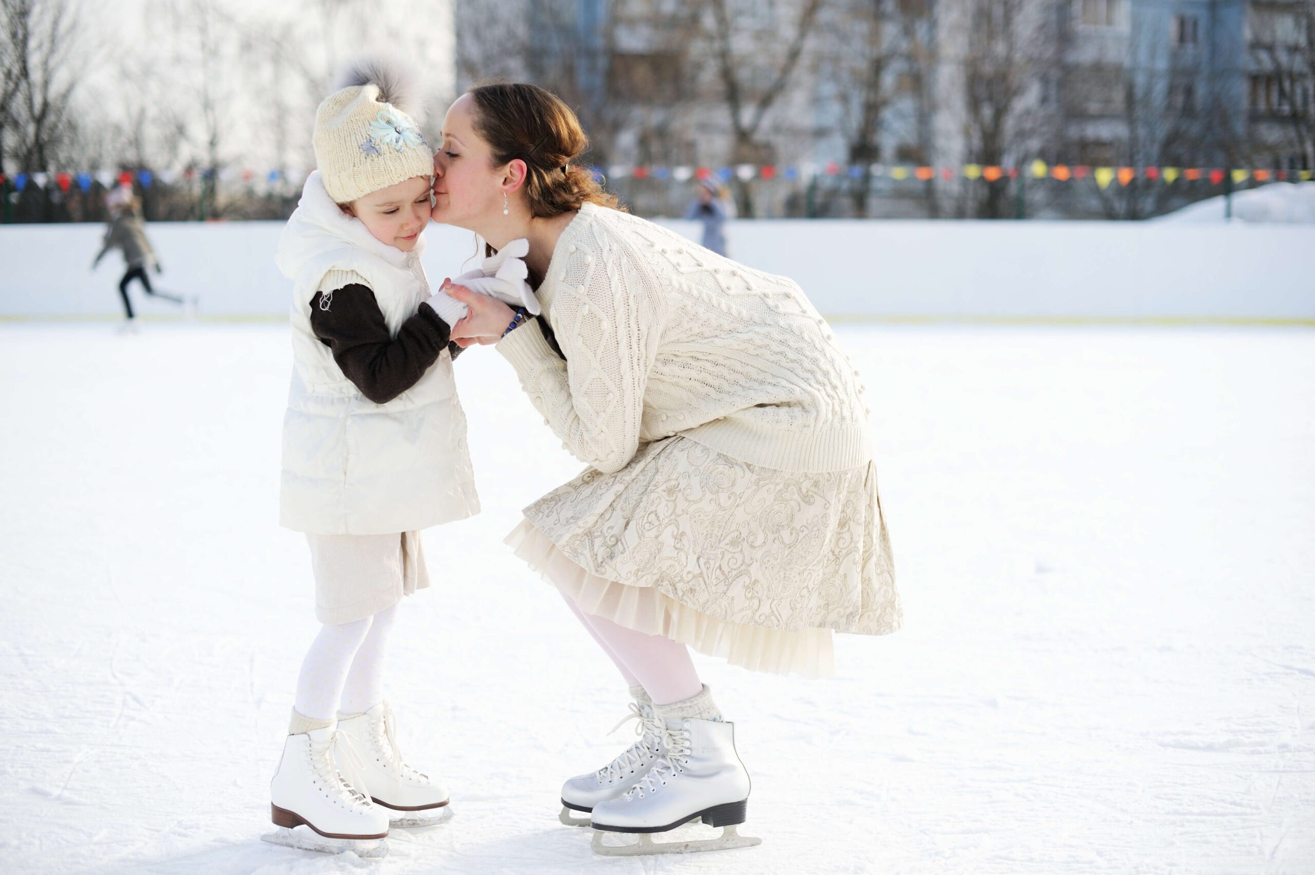 Eine Mutter küsst liebevoll ihre Tochter auf einer Eisbahn. Winterkleidung und Schlittschuhe betonen die Szene. Climate Energy.